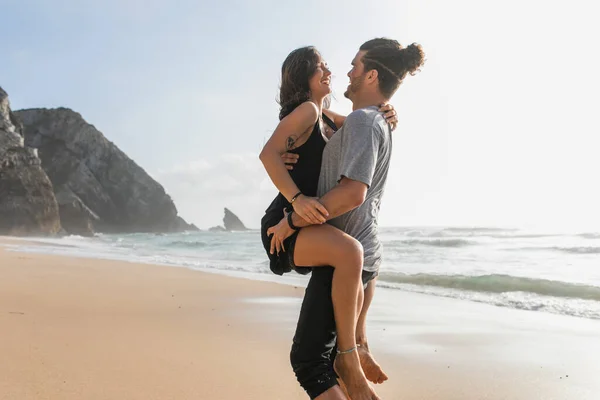 Hombre barbudo feliz levantando mujer tatuada en vestido en la playa cerca del océano - foto de stock