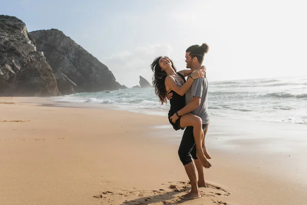 Barbudo hombre levantando feliz novia en vestido en la playa cerca del océano - foto de stock