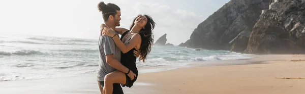 Bearded man lifting happy girlfriend in dress on beach near ocean, banner — Stock Photo