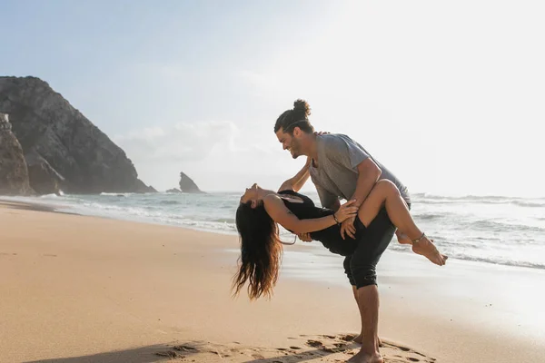 Вид збоку щасливого бородатого чоловіка, який піднімає татуйовану жінку в одязі на пляжі біля океану — стокове фото