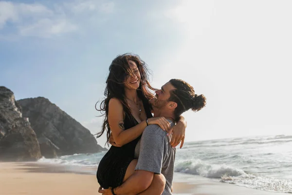 Весёлый бородатый мужчина поднимает татуировку и счастливую женщину в платье на пляже возле океана — стоковое фото