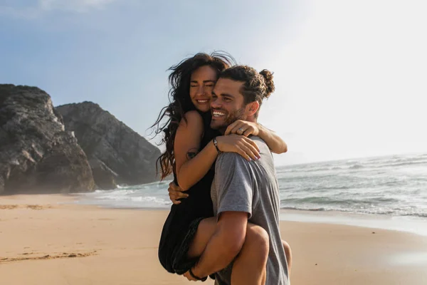 Cheerful man lifting and hugging tattooed woman in dress on beach near ocean — Stock Photo