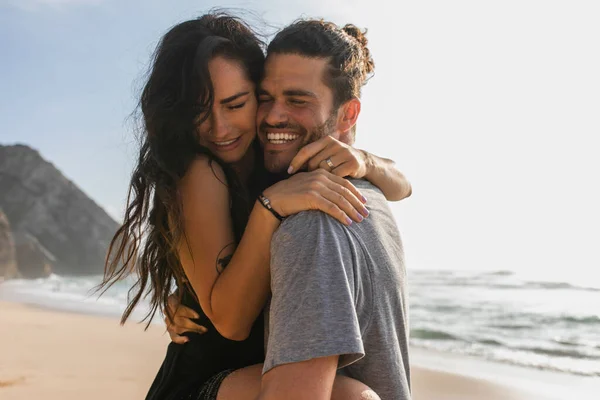 Homem alegre levantando e abraçando mulher tatuada no vestido na praia perto do oceano — Fotografia de Stock