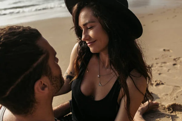 Happy and tattooed woman in hat smiling while looking at boyfriend — Stock Photo