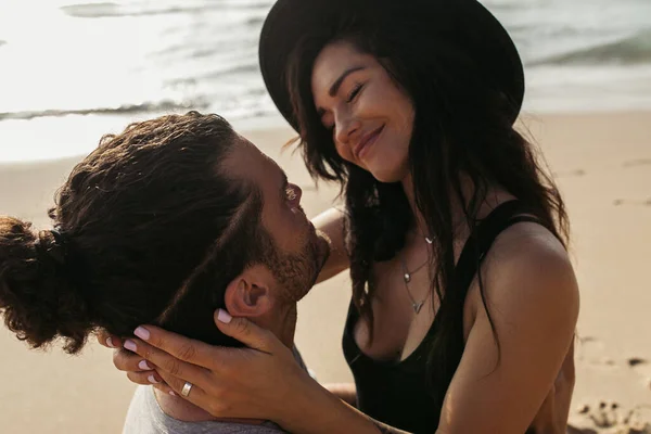 Mulher feliz no chapéu sorrindo enquanto olha para namorado barbudo na praia — Fotografia de Stock