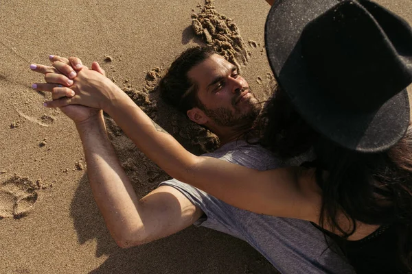 Сверху вид бородатого мужчины, держащегося за руки с татуированной девушкой в шляпе и лежащего на песке — стоковое фото