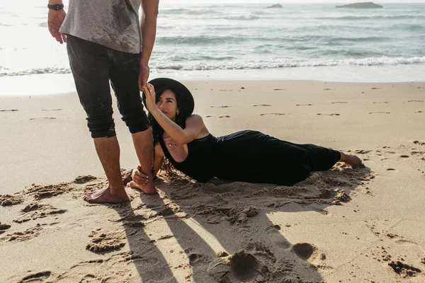 Woman in dress and hat lying on wet sand and hugging leg of man near ocean — Stock Photo