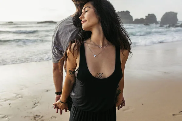 Tattooed woman and man standing back to back and holding hands near ocean — Stock Photo