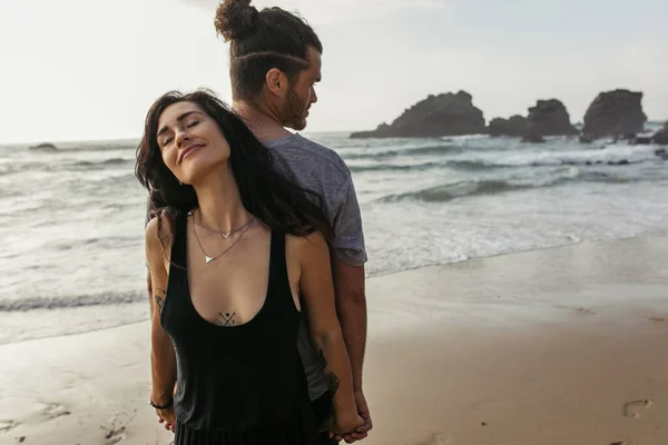 Tattooed woman and bearded man standing back to back and holding hands near ocean — Stock Photo