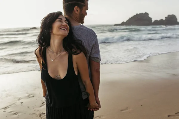 Happy woman and bearded man standing back to back and holding hands near ocean — Stock Photo