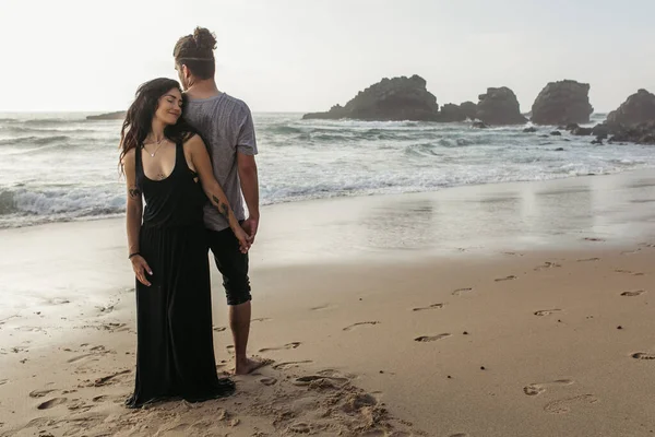 Full length of happy woman in dress and man standing on beach and holding hands near ocean — Stock Photo