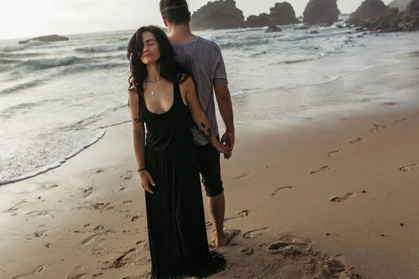 Full length of tattooed woman in dress and man standing on beach and holding hands near ocean — Stock Photo