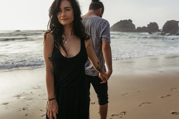 Татуированная женщина в платье и мужчина, держащийся за руки возле океана на пляже в Португалии — стоковое фото