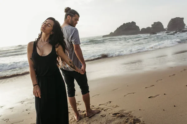 Homme barbu et femme tatouée en robe debout sur la plage et tenant la main près de l'océan — Photo de stock