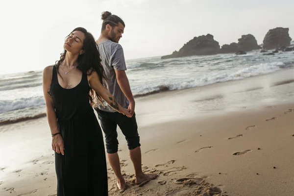 Bearded man and tattooed woman in dress standing on beach and holding hands near ocean — Stock Photo