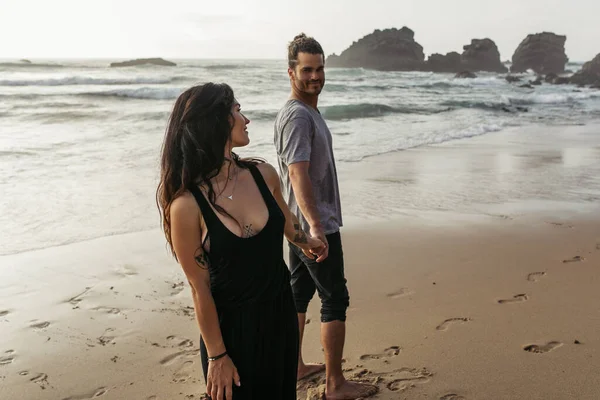 Бородатый мужчина и татуированная женщина в платье улыбаются друг другу на пляже и держатся за руки возле океана — стоковое фото