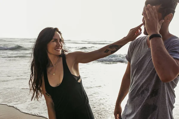 Cheerful and tattooed woman pointing with finger at nose of boyfriend near ocean — Stock Photo