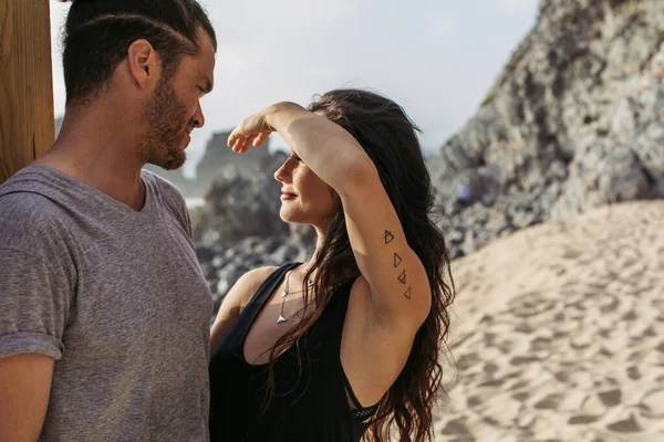 Tattooed woman covering face with hand from sunshine near bearded boyfriend on beach in portugal — Stock Photo