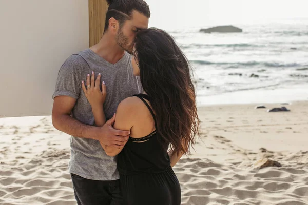 Bearded man kissing brunette girlfriend with wavy hair on beach near ocean — Stock Photo