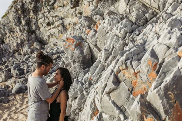 Vista lateral del hombre barbudo abrazándose y mirando a su novia cerca de las rocas en la playa - foto de stock