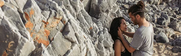 Side view of bearded man hugging and looking at girlfriend near rocks on beach, banner — Stock Photo