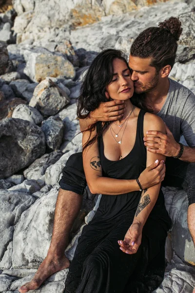 Бородатый мужчина целует татуированную женщину в платье рядом со скалами на пляже — стоковое фото