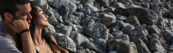 Bärtiger Mann umarmt tätowierte Frau mit geschlossenen Augen in der Nähe von Felsen am Strand, Banner — Stockfoto