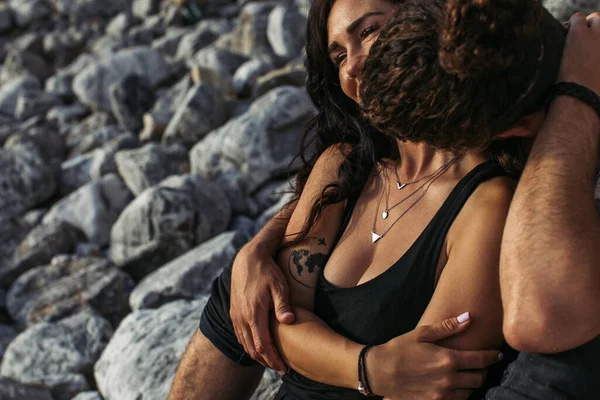 Man kissing tattooed girlfriend in dress near rocks on beach — Stock Photo
