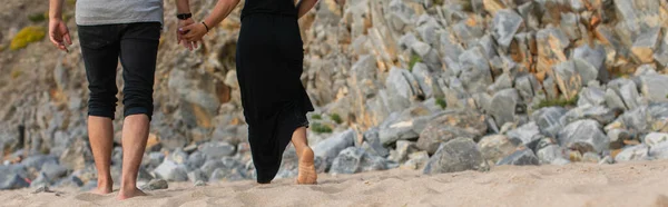 Vue recadrée du couple tenant la main et marchant sur le sable près des rochers, bannière — Photo de stock