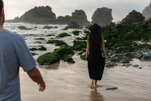 Vista trasera del hombre borroso caminando detrás de la mujer tatuada en vestido negro cerca de musgo verde en piedras en el océano - foto de stock