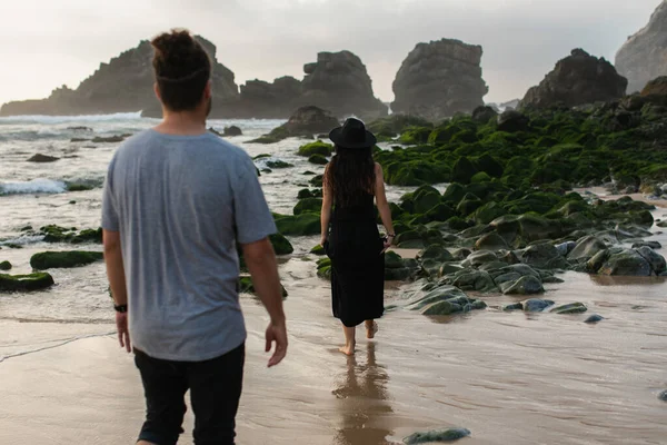 Visão traseira do homem andando atrás de mulher tatuada em vestido preto perto de musgo verde em pedras e oceano — Fotografia de Stock