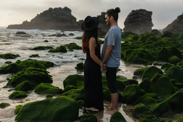 Woman in hat and dress holding hands with happy boyfriend near mossy stones and ocean — Stock Photo