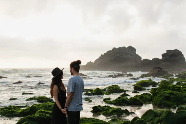 Mulher de chapéu e vestido de mãos dadas com namorado barbudo perto de pedras musgosas e oceano — Fotografia de Stock