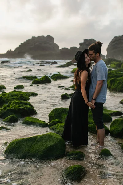 Barbudo hombre besos mejilla de novia en sombrero y vestido cerca musgoso piedras y océano - foto de stock