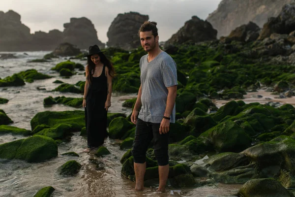 Homem barbudo e mulher tatuada em vestido e chapéu andando perto de pedras musgosas e oceano — Fotografia de Stock