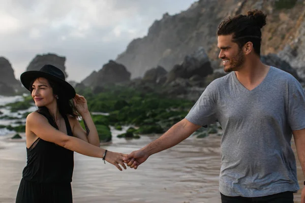 Pleased woman in hat and dress holding hands with bearded boyfriend near ocean — Stock Photo