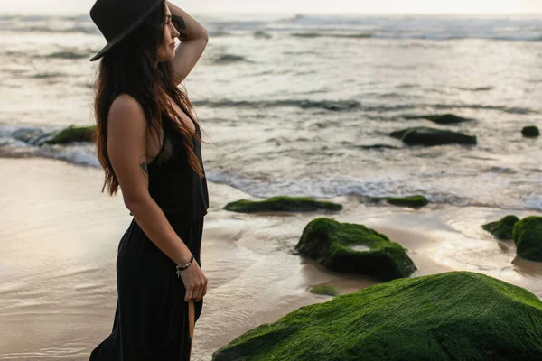 Vista panorámica de mujer tatuada en vestido negro y sombrero mirando al océano cerca de piedras verdes musgosas - foto de stock