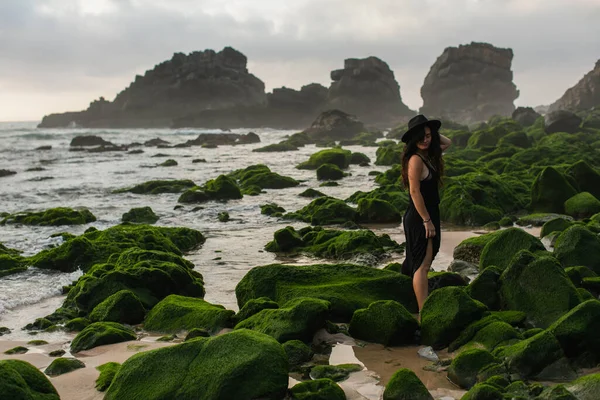 Vista cênica de mulher tatuada em vestido e chapéu em pé perto de pedras verdes musgosas no oceano — Fotografia de Stock
