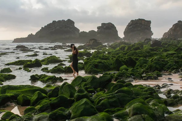 Scenic view of woman in black dress and hat walking near mossy green stones in ocean — Stock Photo