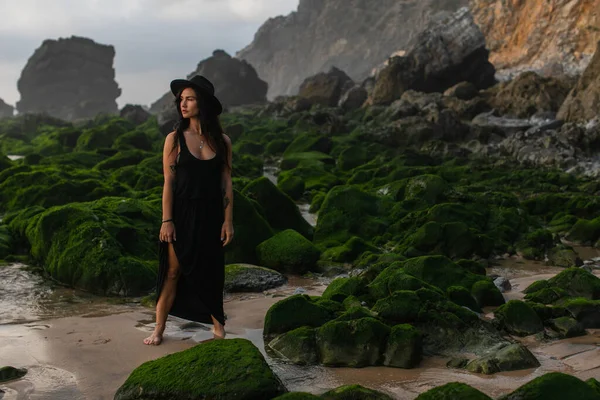 Comprimento total de mulher tatuada em vestido preto e chapéu em pé perto de pedras verdes musgosas — Fotografia de Stock