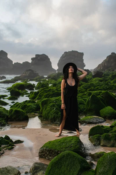 Tattooed woman in black dress and hat standing and looking up near mossy green stones in ocean — Stock Photo