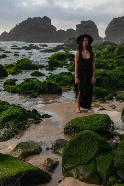 Tattooed woman in black dress and hat standing near mossy green stones in ocean — Stock Photo