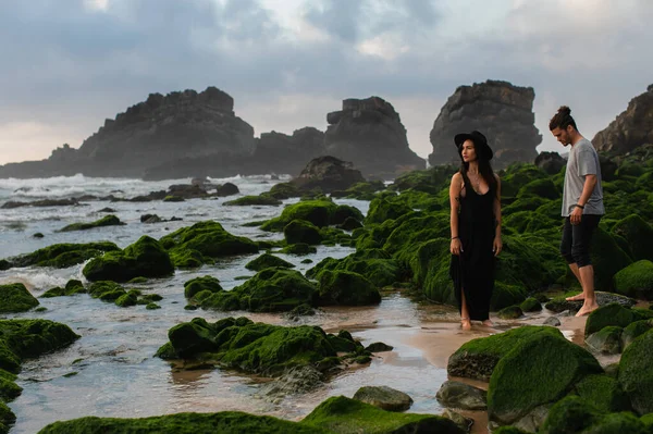 Tattooed woman in hat and dress standing near bearded man and mossy stones in ocean — Stock Photo