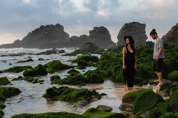 Tätowierte Frau mit Hut und Kleid steht neben bärtigem Mann und bemoosten Steinen im Meer — Stockfoto