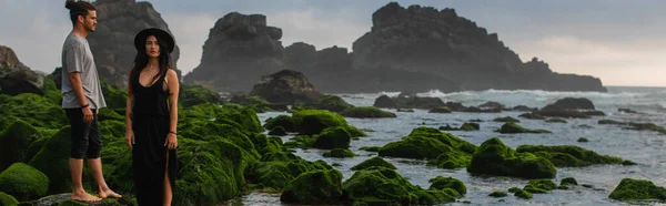 Tätowierte Frau mit Hut und Kleid neben bärtigem Freund und bemoosten Steinen im Meer, Banner — Stockfoto