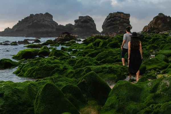 Back view of tattooed woman in hat and dress standing behind man on mossy stones near ocean — Stock Photo