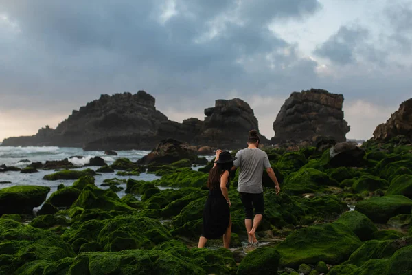 Back view of tattooed woman in hat and dress walking behind man on mossy stones near ocean — Stock Photo