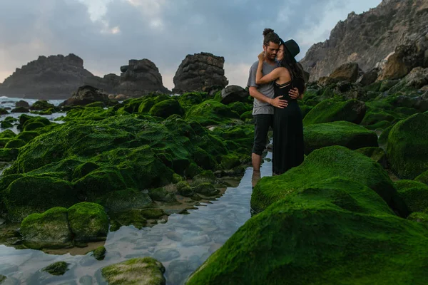 Tattooed woman in hat and dress hugging bearded boyfriend near mossy stones in ocean — Stock Photo