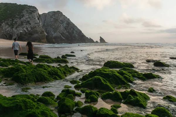 Visão traseira do casal andando perto de pedras musgosas verdes no oceano durante o pôr do sol — Fotografia de Stock