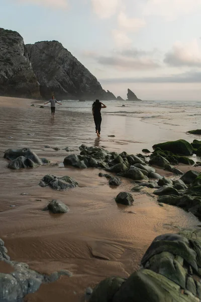 Homem com as mãos estendidas encontrar namorada em chapéu e vestido andando na areia molhada no oceano — Fotografia de Stock